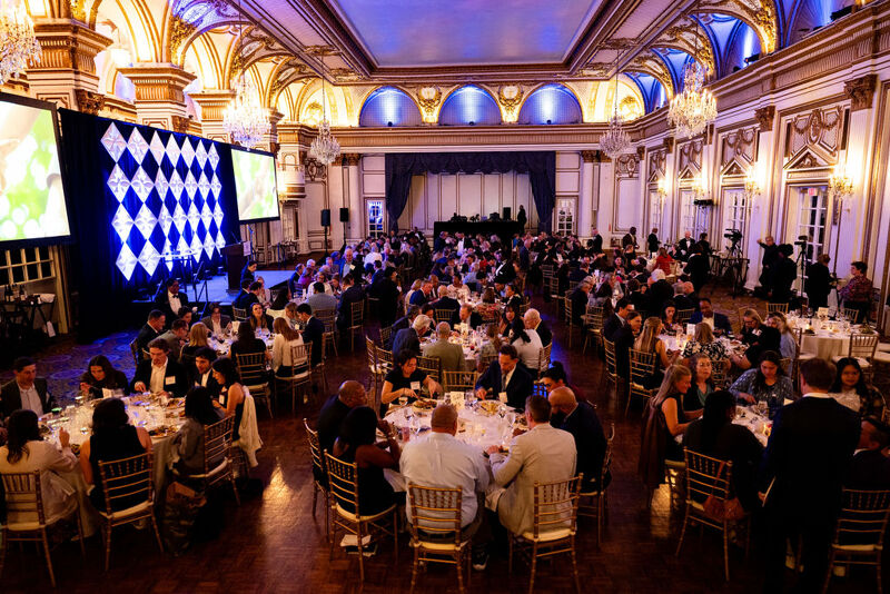 The image shows a large event or gala held in a grand ballroom. Numerous people are seated at round tables, suggesting a formal dinner or awards ceremony. A large screen displays a pattern, and the room is ornately decorated with chandeliers and architectural details. The atmosphere seems lively and celebratory.